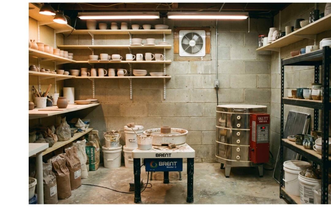 Well-organized home pottery studio in a garage with a pottery wheel, electric kiln, clay storage, and tool shelves