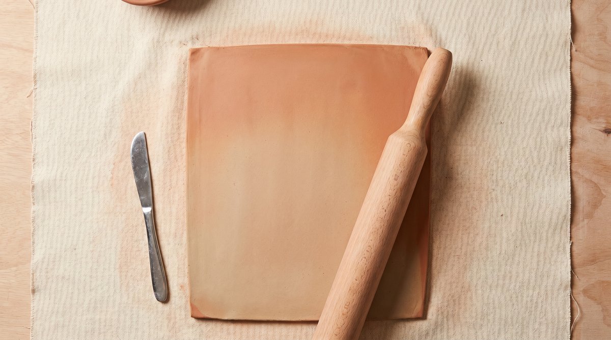 Slab Pottery Technique Setup - Rolling Pin and Clay Slab Clay slab rolled flat on canvas with rolling pin and fettling knife, showing the slab pottery technique setup